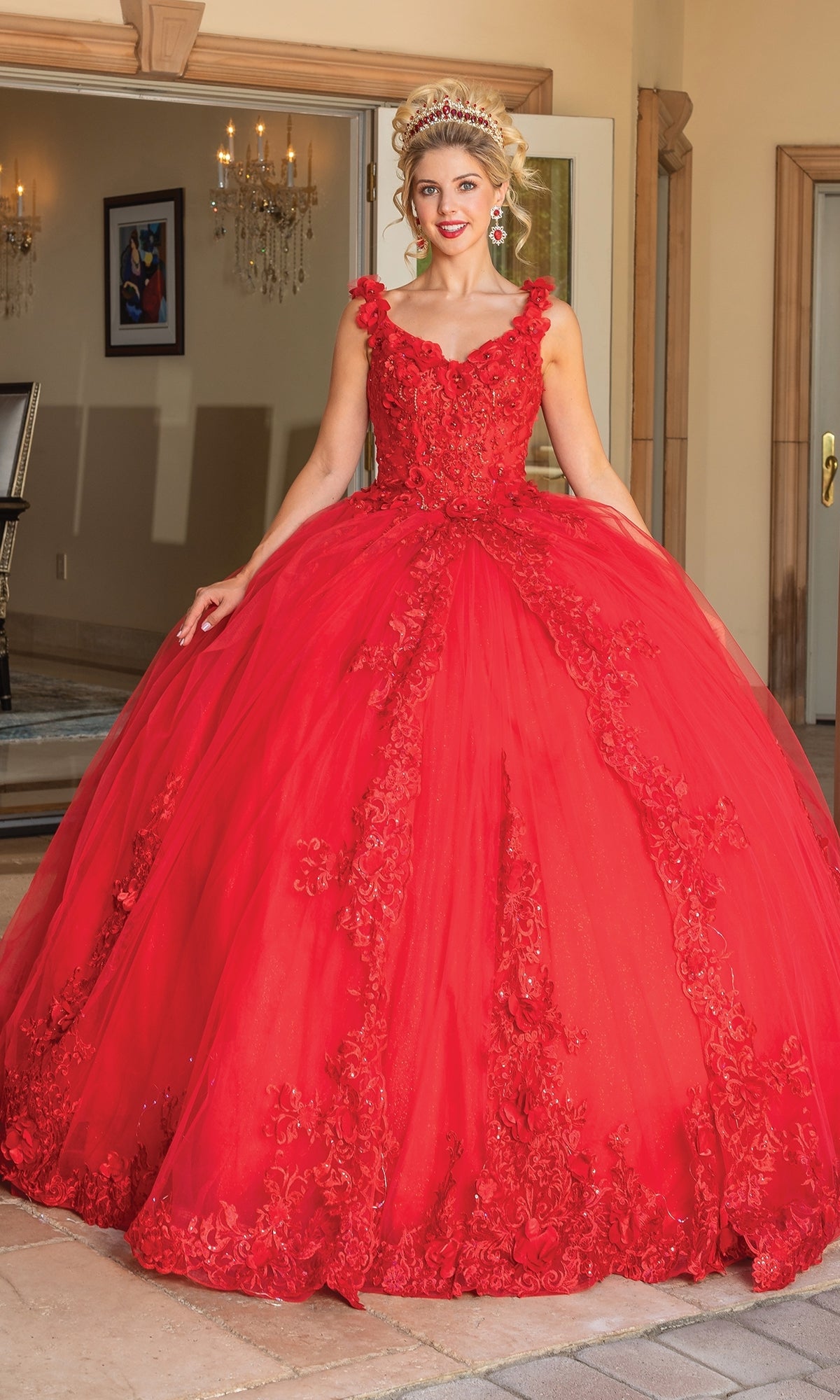 A woman outdoors smiles at the camera, wearing the Quinceanera Dress 1678 By Dancing Queen—a bright red ball gown with intricate embroidery, floral accents, and removable draped sleeves on its full skirt.
